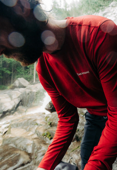 A man wearing a red long-sleeve performance shirt with Montane logo printed on the chest, bent over near a rocky waterfall in a forest.