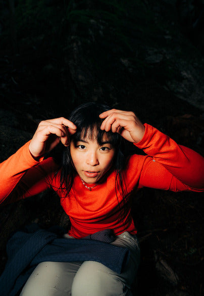 A woman in a bright orange-red long-sleeve top and light grey pants sits in a dark outdoor setting.