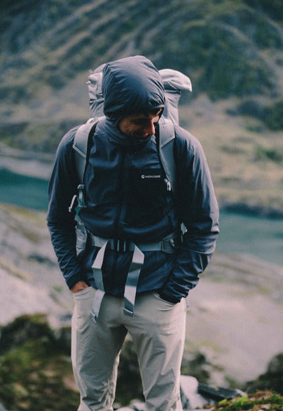 A man wearing a dark blue hooded technical jacket, light gray hiking pants, and a large gray backpack stands outdoors in a mountainous, misty environment.