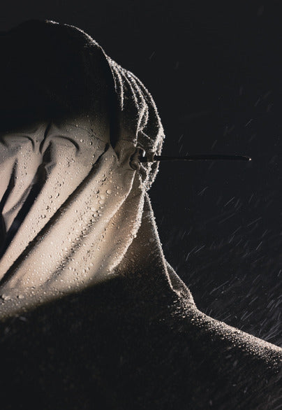 A dramatic close-up of a hooded figure in dark rain, with water droplets glistening on the fabric